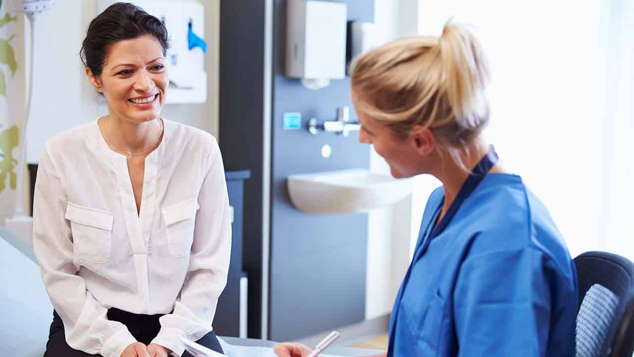 a female client is smiling while talking to a physician assistant who is wear blue scrubs a stethoscope is around her neck